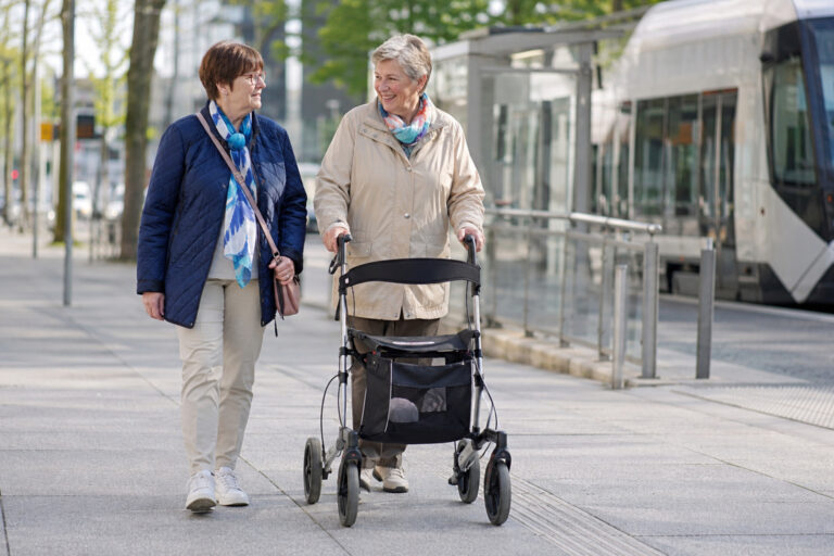 Ältere Frau mit Rollator ist gemeinsam mit einer Begleitperson im öffentlichen Raum unterwegs.